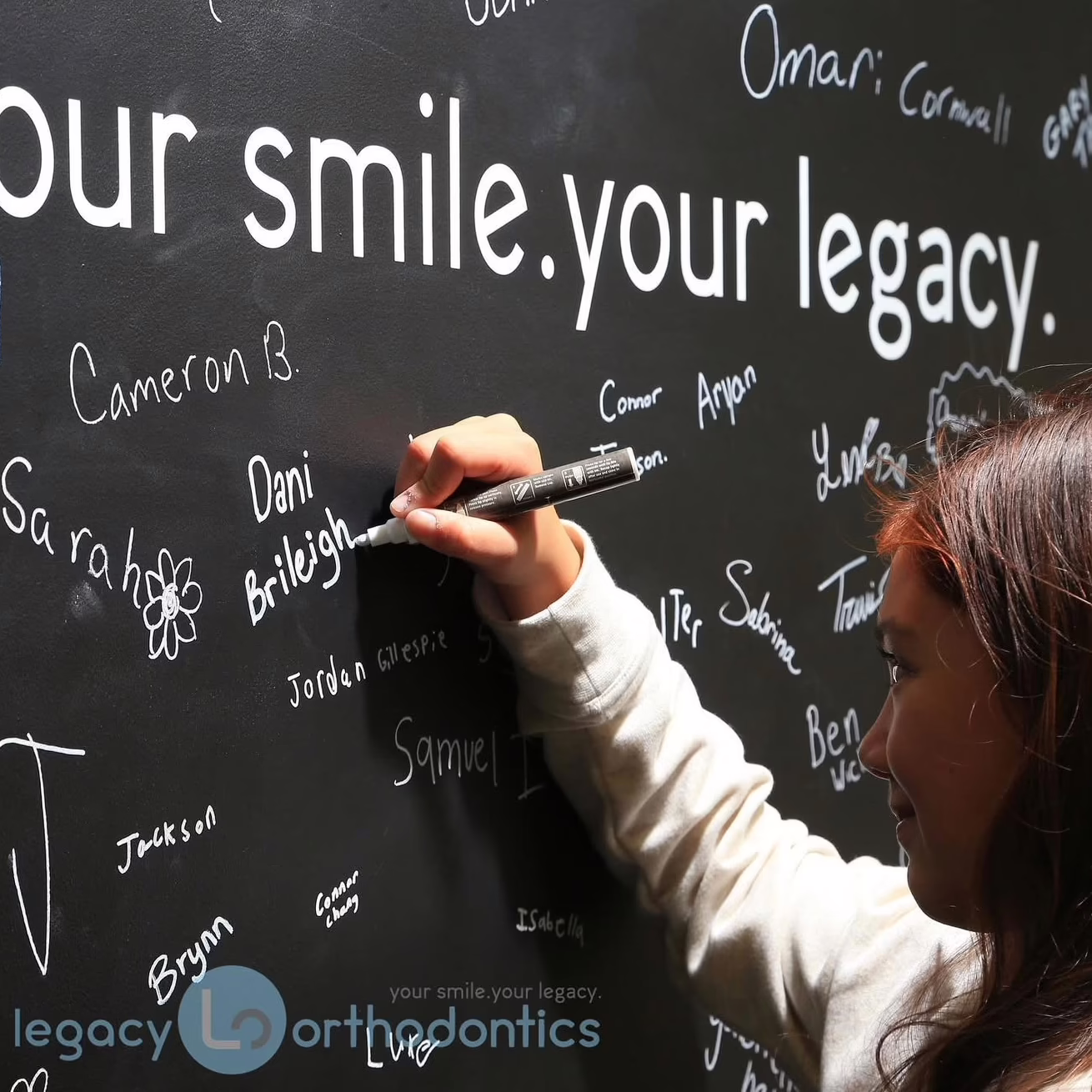 Girl writing on a black wall with the text "your smile. your legacy." and various names, representing Legacy Orthodontics' community engagement and personalized orthodontic care in Leesburg.